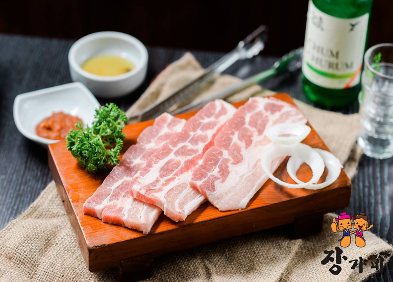 Three slices of samgyupsal laid on a board with Soju and dipping sauces in the background